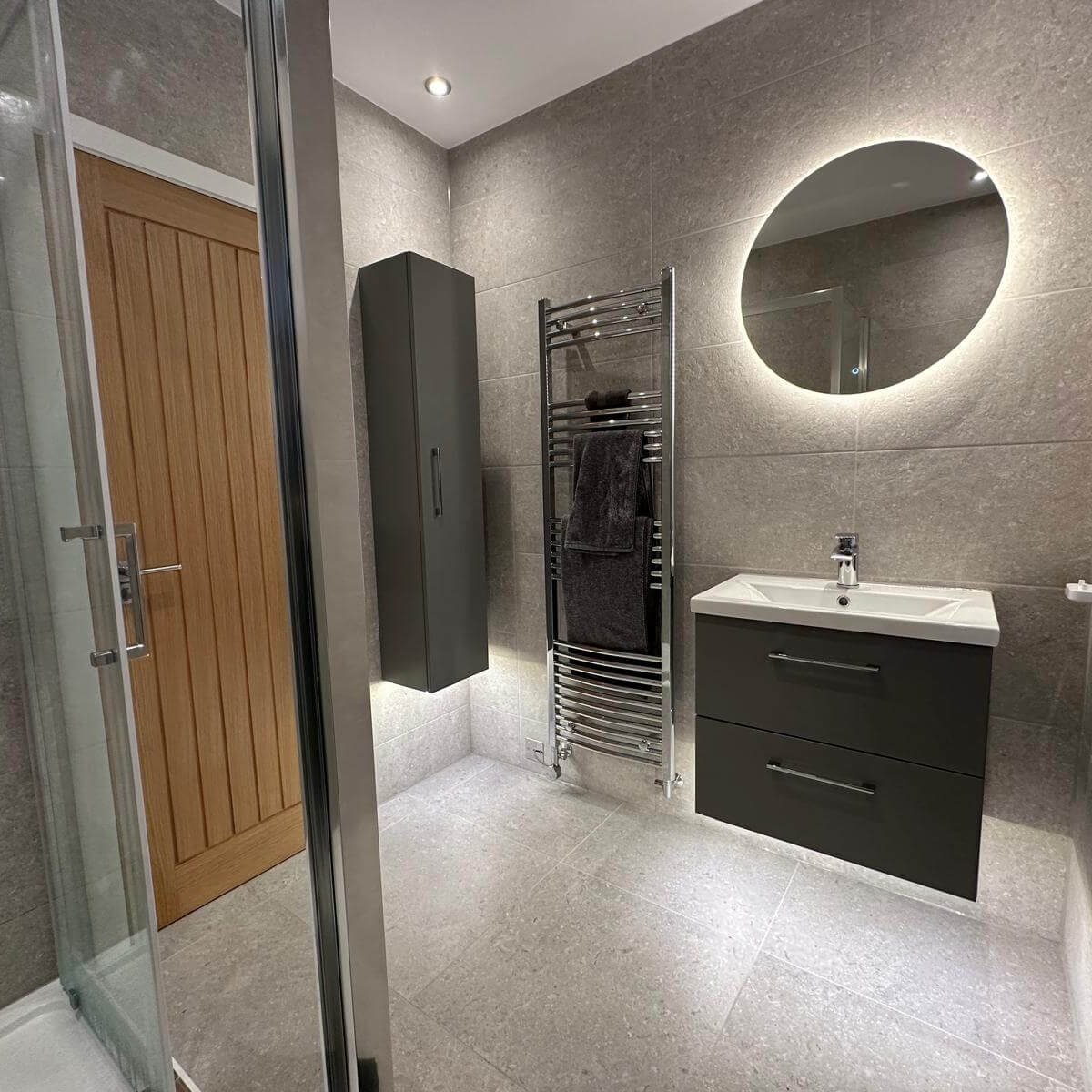 Grey tiled bathroom with a wall-hung vanity, illuminated round mirror and heated towel rail in Brislington