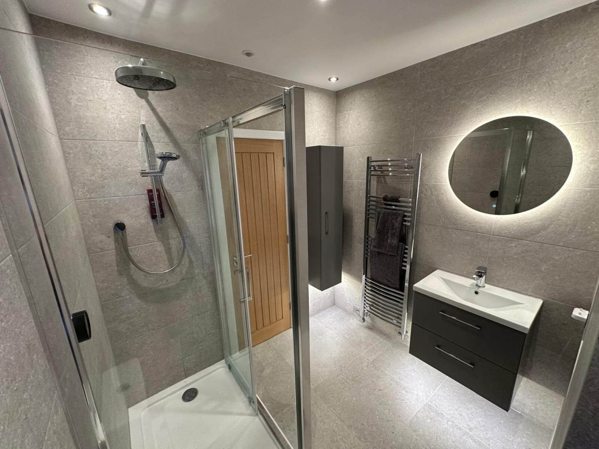 Glass shower enclosure beside a grey tiled vanity and illuminated mirror